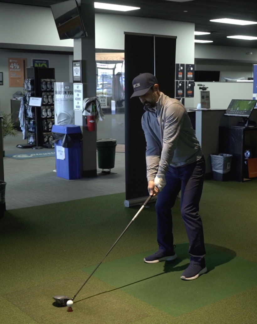 golf fit conclusion - Tour Quality Golf | Indoor Lessons, Fittings & Virtual Golf Man in a cap practicing golf indoors, preparing to swing a club from a mat with a golf ball on a tee.