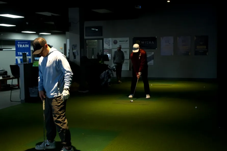 club-repair-pic-890 - Tour Quality Golf | Indoor Lessons, Fittings & Virtual Golf Two golfers practice indoors on artificial turf; one in a light hoodie prepares a swing, another tees off in the background.
