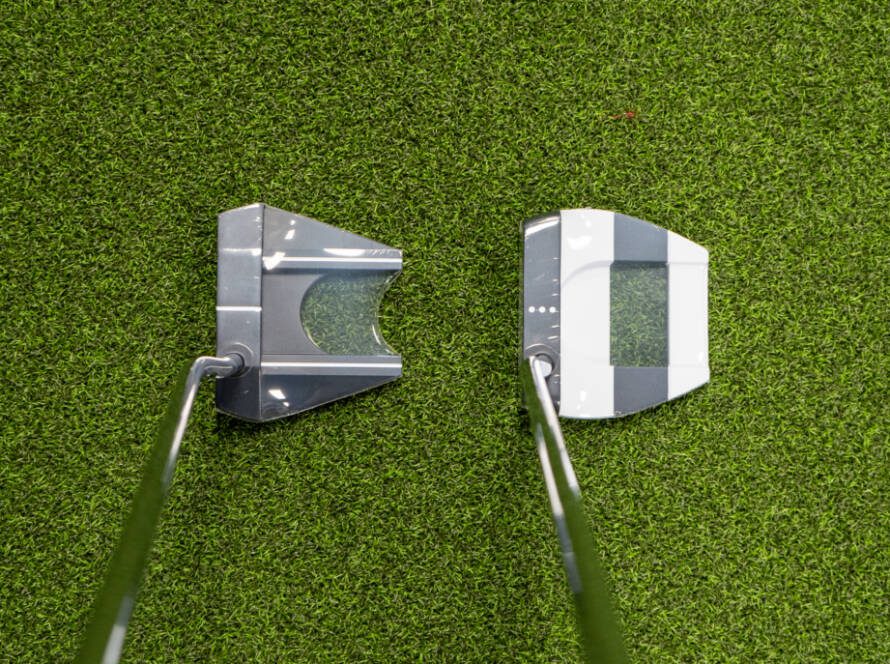 Two golf putter heads with shafts rest on green artificial turf, facing each other from above.